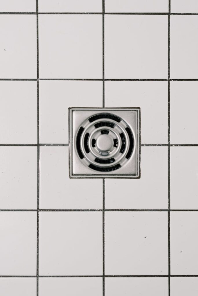 Photo by Ksenia Chernaya Top view of white ceramic tile floor with stainless steel square shaped cover of drain in contemporary shower